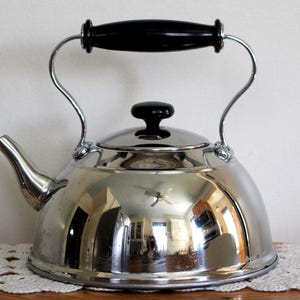 May include: A shiny, stainless steel tea kettle with a black handle and lid knob. The kettle has a curved spout and a reflective surface, showing the surrounding room. The handle is arched and attached to the body of the kettle.