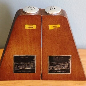 May include: Wooden salt and pepper shakers with a brown finish. Each shaker has a silver top with holes and a yellow letter, "S" and "P". Black labels display a cable car illustration and "San Francisco".