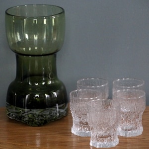 May include: A set of vintage glassware, including a large olive green glass vase and five clear textured shot glasses. The vase has a wide top and a textured base. The shot glasses have a textured, bark-like appearance.