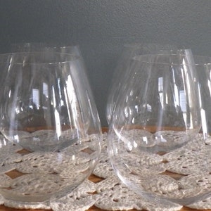 May include: A collection of clear, stemless wine glasses. The glasses have a rounded bowl shape and are arranged on a white, crocheted doily. The background is a muted gray, providing a neutral backdrop for the glassware.