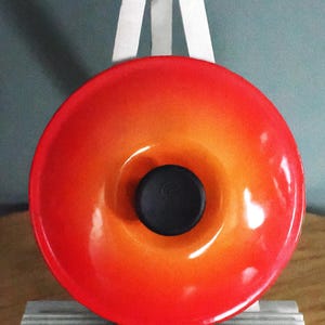 Pode incluir: Uma vibrante tampa redonda de panela de ferro fundido esmaltado laranja e vermelho, com um botão preto no centro. A tampa é exibida em um cavalete branco contra um fundo verde-azulado suave. A tampa tem um acabamento brilhante.