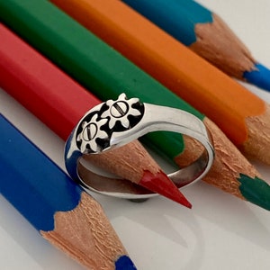 May include: A silver-toned metal ring featuring two interlocking gear-shaped designs on the top surface. Each gear has a central horizontal line detail. The ring is positioned against a background of several colored wooden pencils.