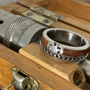 May include: A silver-toned metal ring featuring a dark brown wood inlay and a gear-shaped accent on the outer band. The ring is positioned next to a metal micrometer tool with visible measurement markings, showing a scale from 30 to 40.