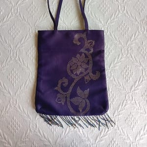 May include: A purple tote bag with a beaded fringe and a silver floral design. The design is made of small, shiny beads.