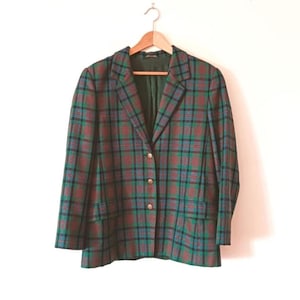 Op de afbeelding: Een vintage geruite blazer in groen, rood en blauw, met goudkleurige knopen. De blazer hangt aan een houten hanger tegen een witte achtergrond. De jas heeft twee voorzakken.