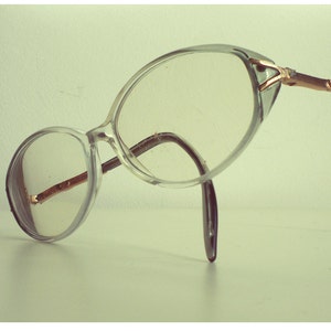 May include: Clear plastic framed glasses with a gold metal bridge and arms. The glasses are lying on a white surface.