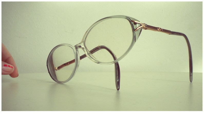 May include: Clear plastic framed glasses with a gold metal bridge and arms. The glasses are lying on a white surface.