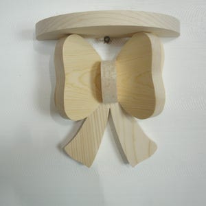 May include: A wooden wall shelf in the shape of a bow. The shelf has a curved top and a bow-shaped design below, with two large loops and two hanging ribbons. The wood has a natural, light color.