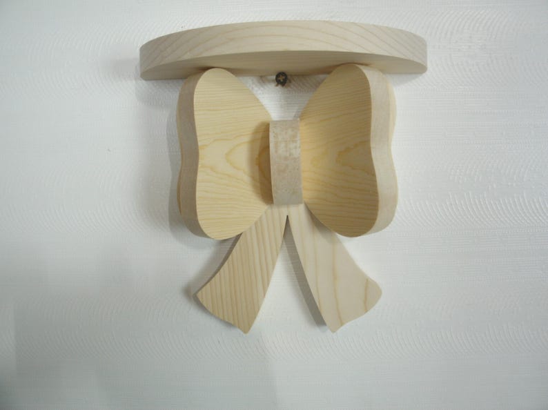 May include: A wooden wall shelf in the shape of a bow. The shelf has a curved top and a bow-shaped design below, with two large loops and two hanging ribbons. The wood has a natural, light color.