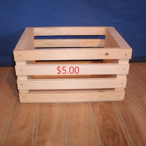 May include: A natural wood crate with a price tag of $5.00. The crate is made of unfinished wood and has a rectangular shape with four sides and a bottom. The crate has four horizontal slats on the front and back sides.