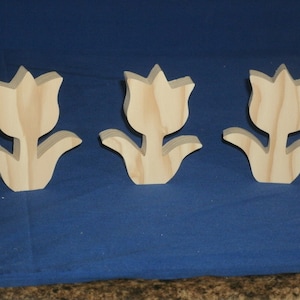 Peut inclure: Trois formes de tulipes en bois non fini, chacune avec une seule tige et deux feuilles. Les tulipes sont disposées en rangée sur une surface bleue.