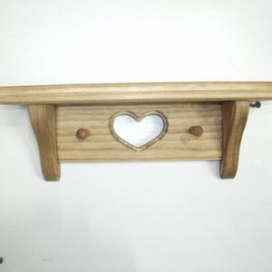 May include: Wooden shelf with a heart-shaped cutout and two small pegs. The shelf is made of light-colored wood and has a simple, rustic design. The shelf is approximately 18 inches wide.