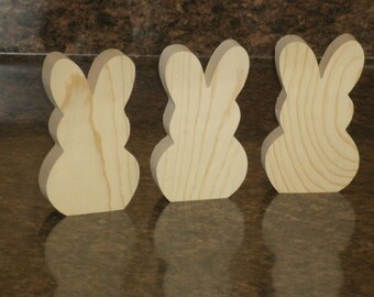 May include: Three unfinished wooden bunny shapes, each with a rounded body and two ears. The bunnies are arranged in a row from left to right.
