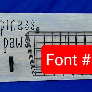 Puede incluir: Un letrero de madera blanco con el texto "Happiness has paws" en letras negras. El letrero tiene dos ganchos negros y una cesta de alambre negro. El texto "Font #3" est&aacute; escrito en blanco sobre un fondo rojo.