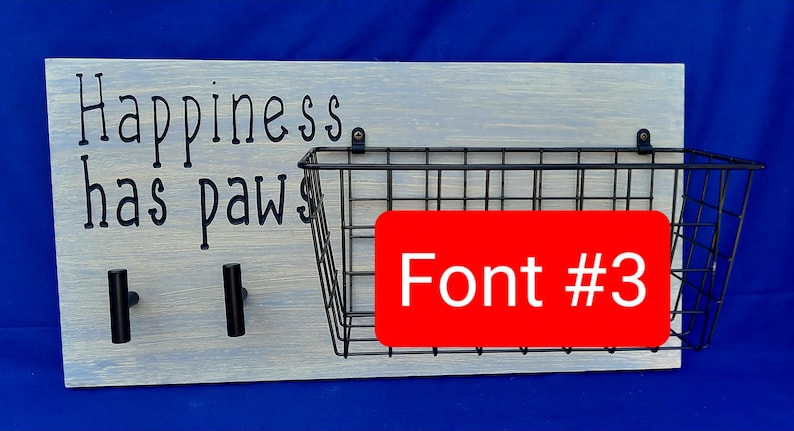 Puede incluir: Un letrero de madera blanco con el texto "Happiness has paws" en letras negras. El letrero tiene dos ganchos negros y una cesta de alambre negro. El texto "Font #3" est&aacute; escrito en blanco sobre un fondo rojo.