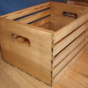 May include: A wooden crate with a natural finish and two handles. The crate has a rectangular shape and is made of wood slats.