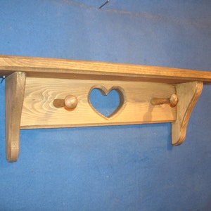 May include: Wooden shelf with a heart-shaped cutout and two wooden pegs. The shelf is made of light brown wood and has a rustic, handmade appearance. The shelf is designed for wall mounting and is suitable for displaying small items.