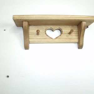 May include: A small, wooden shelf with a heart-shaped cutout and two pegs. The shelf is made of light-colored wood and has a simple, rustic design. The shelf is mounted on a white wall.