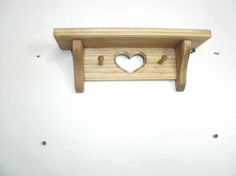 May include: A small, wooden shelf with a heart-shaped cutout and two pegs. The shelf is made of light-colored wood and has a simple, rustic design. The shelf is mounted on a white wall.