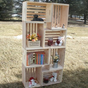 Op de afbeelding: Een houten krattenrek met meerdere planken, elk gevuld met verschillende items, waaronder boeken, kaarsen, knuffels en bloemen. Het rek is gemaakt van natuurlijk hout en heeft een rustieke uitstraling.