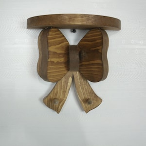May include: A wooden bow-shaped shelf with a dark brown finish. The shelf has a rounded top and two downward-facing bow loops with two pegs. The shelf is mounted on a white wall.