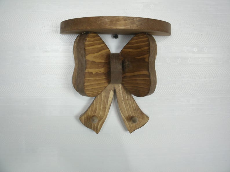 May include: A wooden bow-shaped shelf with a dark brown finish. The shelf has a rounded top and two downward-facing bow loops with two pegs. The shelf is mounted on a white wall.