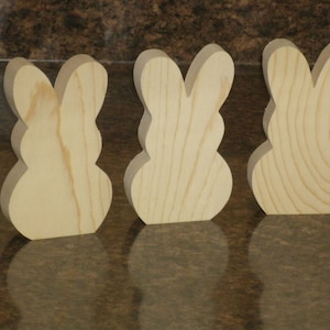 May include: Three unfinished wooden bunny shapes, each with a rounded body and two ears. The bunnies are arranged in a row from left to right.