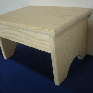 May include: A light-colored wooden step stool featuring a rectangular top surface with rounded edges and four sturdy legs. The stool is constructed from natural wood with visible grain patterns and has arched cutouts at the base of each side.