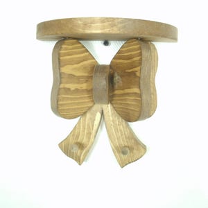 May include: A wooden wall shelf with a bow-shaped design. The shelf is made of light brown wood and features a round top. The bow has a detailed grain pattern, and the shelf is mounted on a white wall.