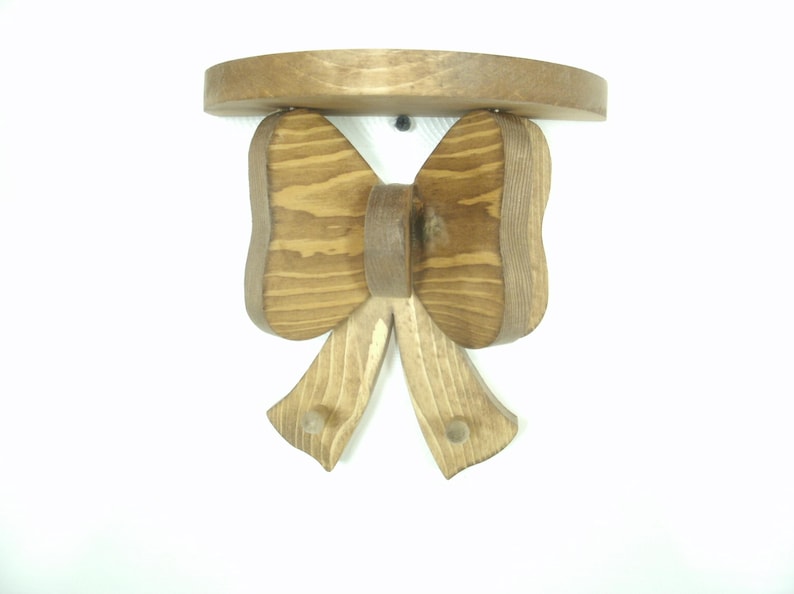 May include: A wooden wall shelf with a bow-shaped design. The shelf is made of light brown wood and features a round top. The bow has a detailed grain pattern, and the shelf is mounted on a white wall.