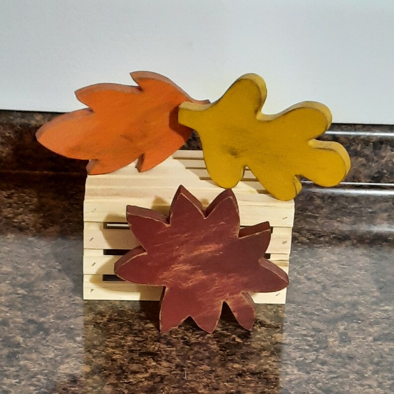 May include: Three wooden fall-themed decorations: an orange leaf, a yellow leaf, and a burgundy starburst shape. They rest on a small, light-colored wooden crate. The items have a rustic, distressed finish.