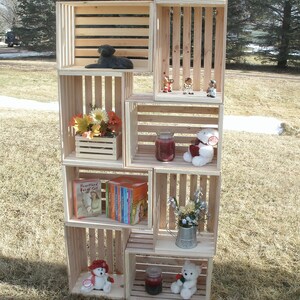 Op de afbeelding: Een houten krattenrek met meerdere planken, elk met verschillende items, waaronder een zwart hondenbeeldje, een boek getiteld "Heartland Boxed Set", een kleine potplant, een witte teddybeer en een klein potje kaarsen.