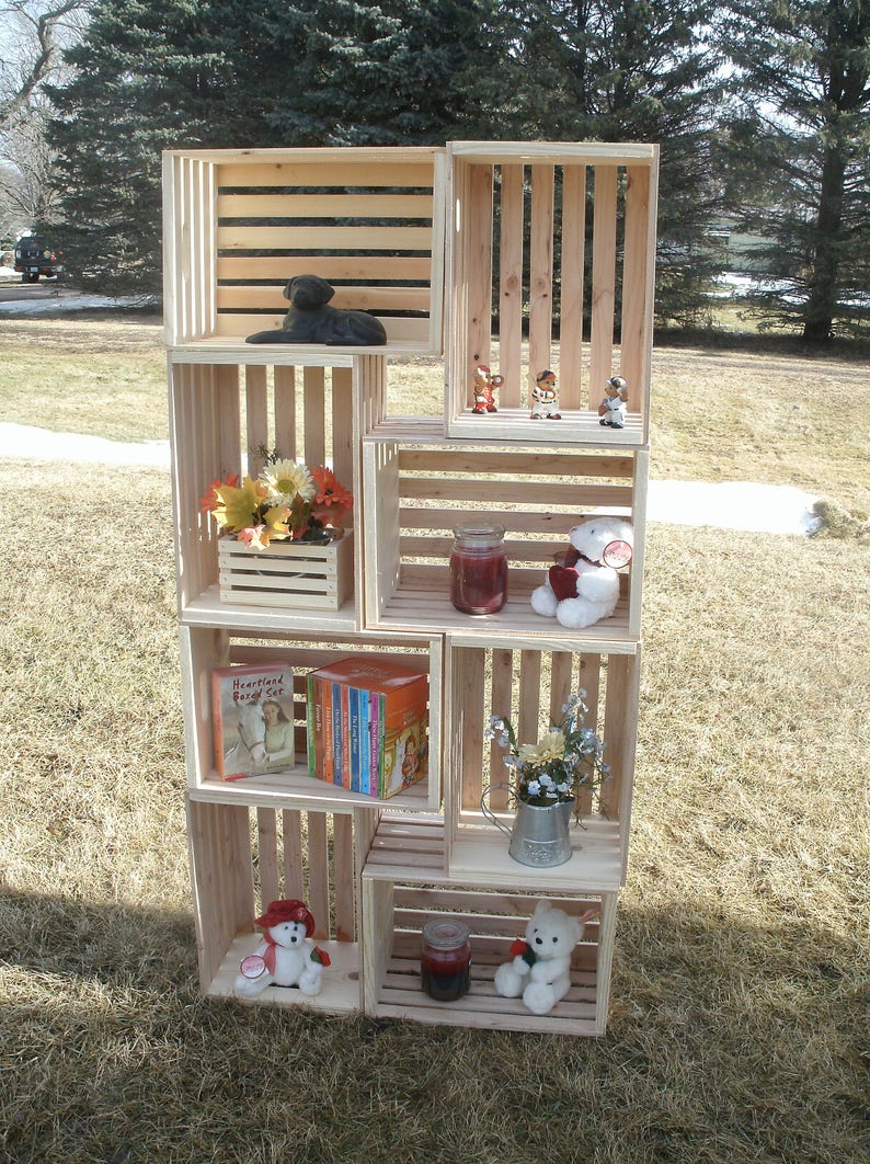 Op de afbeelding: Een houten krattenrek met meerdere planken, elk met verschillende items, waaronder een zwart hondenbeeldje, een boek getiteld "Heartland Boxed Set", een kleine potplant, een witte teddybeer en een klein potje kaarsen.