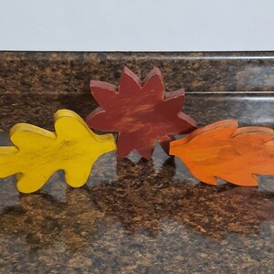 May include: Three wooden fall-themed leaf decorations in yellow, orange, and red. The yellow leaf is on the left, the red flower-shaped leaf is in the middle, and the orange leaf is on the right. The leaves are arranged on a brown surface.