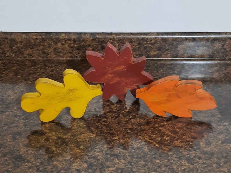 May include: Three wooden fall-themed leaf decorations in yellow, orange, and red. The yellow leaf is on the left, the red flower-shaped leaf is in the middle, and the orange leaf is on the right. The leaves are arranged on a brown surface.
