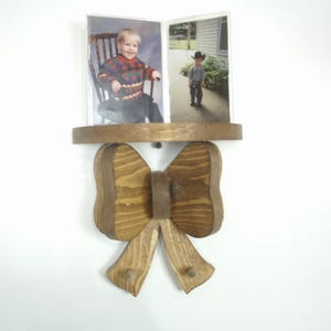 May include: Wooden photo frame in the shape of a bow. The frame holds two photos, one of a child in a chair and another of a child in a cowboy hat. The frame is made of light brown wood and hangs on a white wall.
