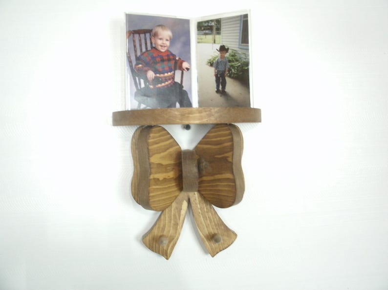May include: Wooden photo frame in the shape of a bow. The frame holds two photos, one of a child in a chair and another of a child in a cowboy hat. The frame is made of light brown wood and hangs on a white wall.