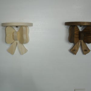 May include: Two wooden bow-shaped shelves. One is light-colored, the other is stained a dark brown. Each shelf has a circular top and a bow-shaped support with two hooks. The shelves are mounted on a white wall.