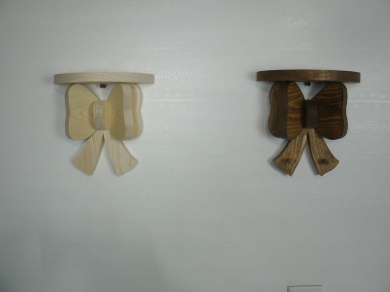 May include: Two wooden bow-shaped shelves. One is light-colored, the other is stained a dark brown. Each shelf has a circular top and a bow-shaped support with two hooks. The shelves are mounted on a white wall.