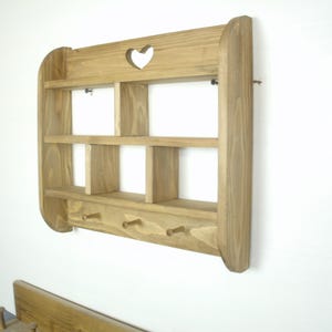 K&ouml;nnte beinhalten: Wandregal aus Holz mit herzf&ouml;rmigem Ausschnitt und drei Haken. Das Regal hat zwei Regalb&ouml;den und mehrere F&auml;cher. Das Holz hat eine nat&uuml;rliche Oberfl&auml;che. Das Regal ist an einer wei&szlig;en Wand befestigt.