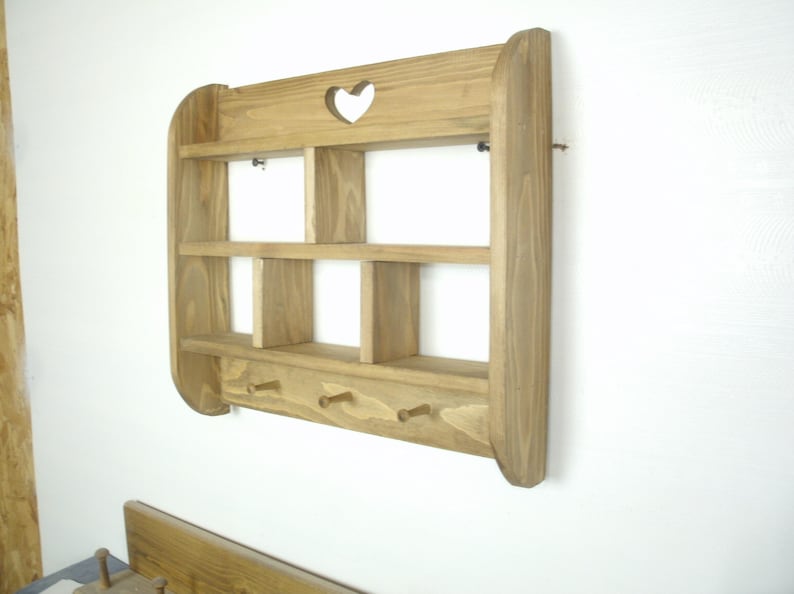 K&ouml;nnte beinhalten: Wandregal aus Holz mit herzf&ouml;rmigem Ausschnitt und drei Haken. Das Regal hat zwei Regalb&ouml;den und mehrere F&auml;cher. Das Holz hat eine nat&uuml;rliche Oberfl&auml;che. Das Regal ist an einer wei&szlig;en Wand befestigt.