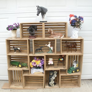 May include: A wooden crate shelf unit with multiple shelves, decorated with various figurines, including horses, a moose, a bear, and a teddy bear. The shelves are filled with flowers and other decorative items. The unit is made of natural wood and has a rustic look.