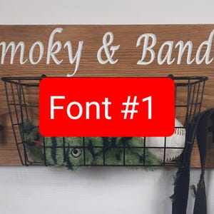 Puede incluir: Un letrero de madera con el texto "Smoky & Bandit" en letras blancas. Una cesta de alambre negro con un juguete de peluche verde y negro y una pelota de b&eacute;isbol blanca cuelga debajo del letrero. El texto "Font #1" est&aacute; escrito en rojo sobre un fondo blanco.