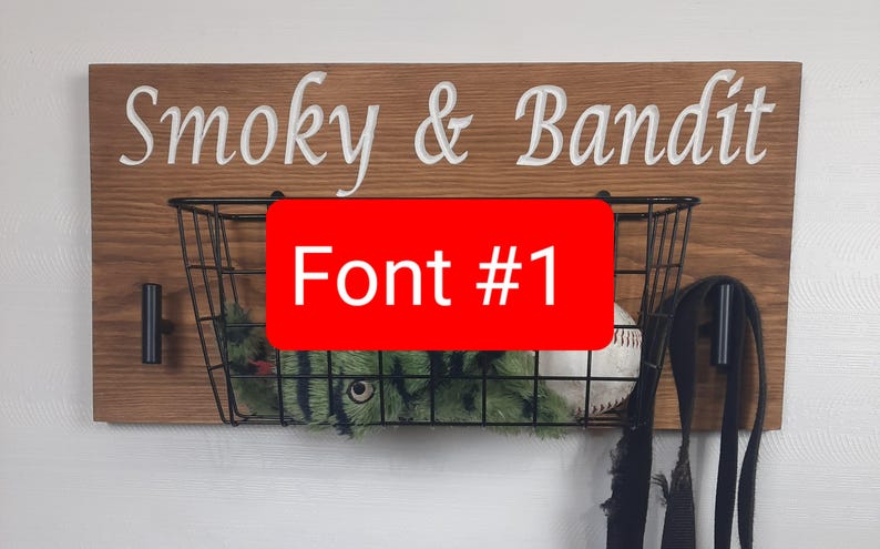 Puede incluir: Un letrero de madera con el texto "Smoky & Bandit" en letras blancas. Una cesta de alambre negro con un juguete de peluche verde y negro y una pelota de b&eacute;isbol blanca cuelga debajo del letrero. El texto "Font #1" est&aacute; escrito en rojo sobre un fondo blanco.