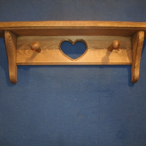 May include: A wooden shelf with a heart-shaped cutout and two wooden knobs. The shelf is made of light-colored wood and has a simple, rustic design. The shelf is mounted on a blue wall and is suitable for displaying small items.