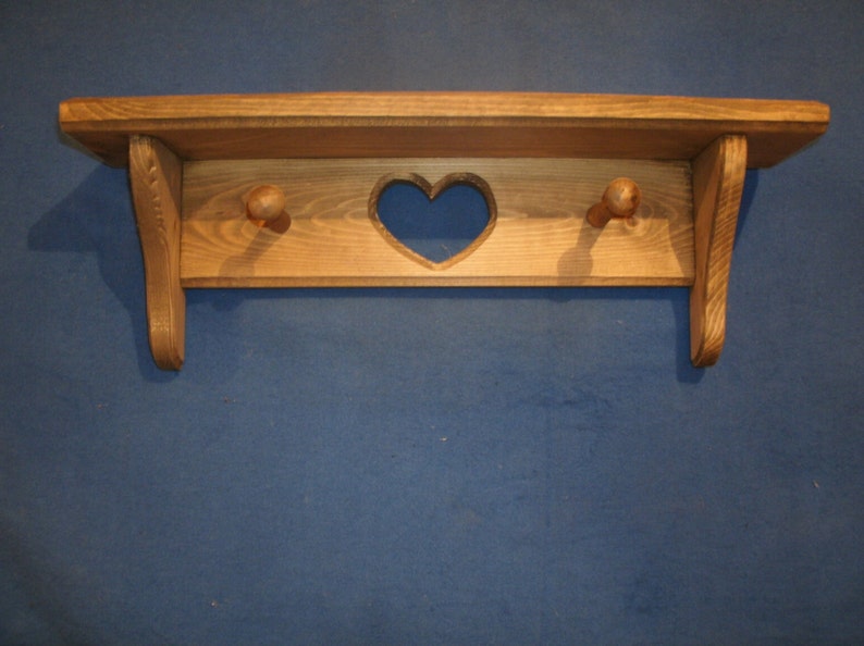 May include: A wooden shelf with a heart-shaped cutout and two wooden knobs. The shelf is made of light-colored wood and has a simple, rustic design. The shelf is mounted on a blue wall and is suitable for displaying small items.