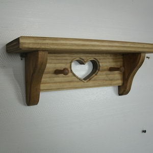 May include: Wooden shelf with a heart-shaped cutout and two pegs. The shelf is made of light-colored wood and has a rustic, country style. The shelf is approximately 12 inches wide.