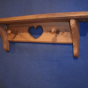 May include: A wooden shelf with a heart-shaped cutout and two wooden pegs. The shelf is made of light brown wood and has a rustic, handmade appearance. The shelf is designed for wall mounting.