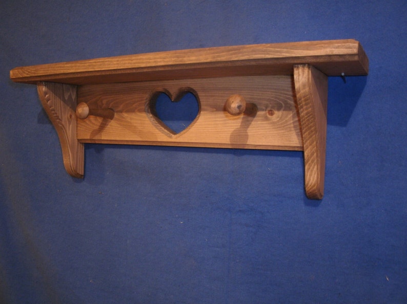 May include: A wooden shelf with a heart-shaped cutout and two wooden pegs. The shelf is made of light brown wood and has a rustic, handmade appearance. The shelf is designed for wall mounting.