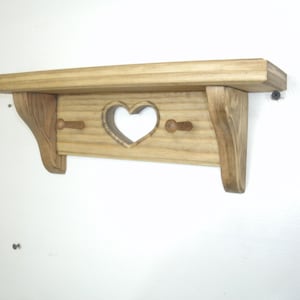 May include: A wooden shelf with a heart-shaped cutout and two pegs below. The shelf is made of light-colored wood with a natural finish. The shelf is mounted on a white wall.